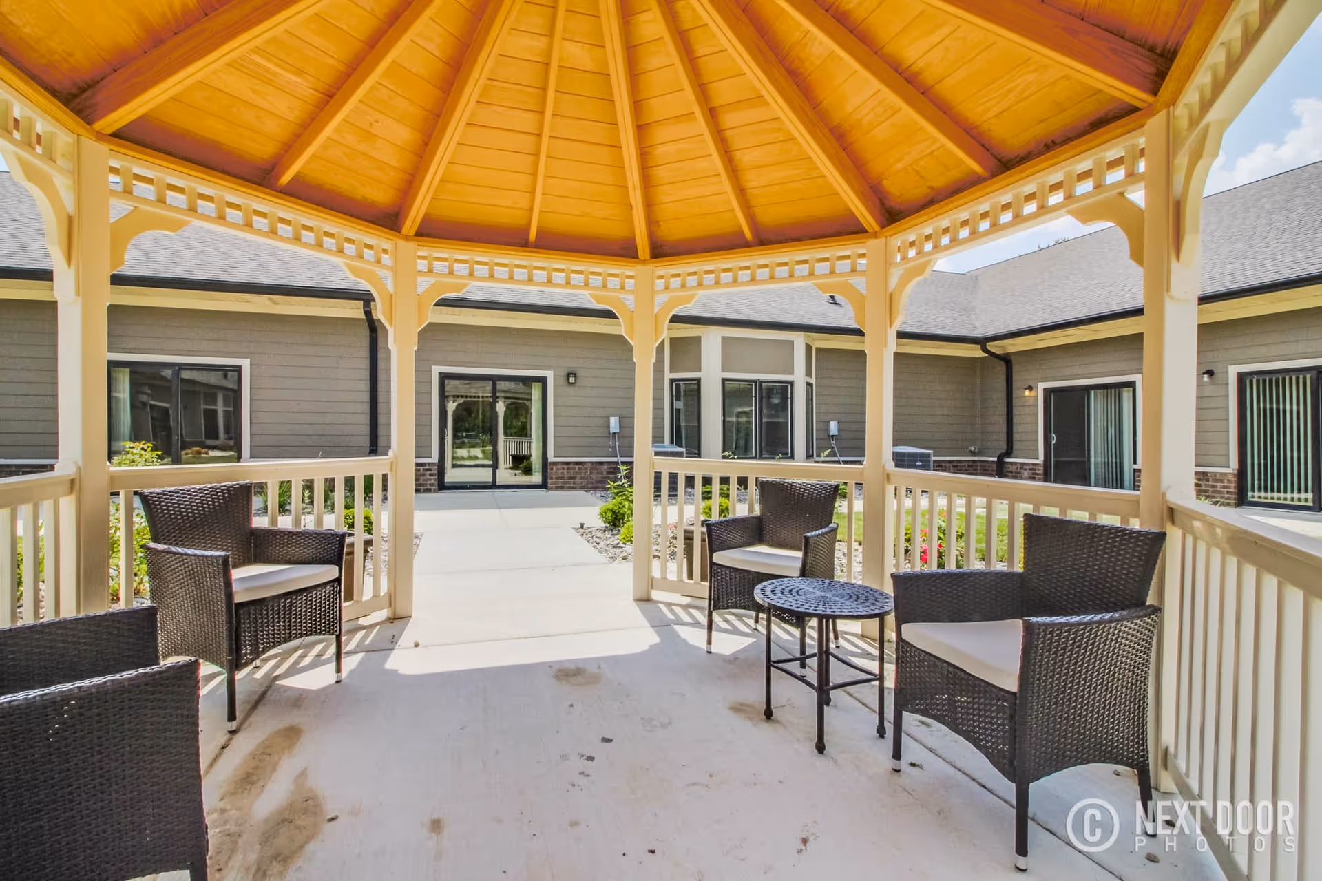 View from inside a wooden gazebo with a peaked roof, furnished with four wicker chairs with cushions and a small round metal table, overlooking a courtyard area of a senior living facility with gray siding and multiple windows and doors.