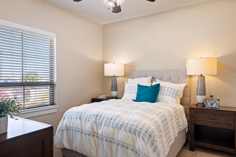 Sunlit bedroom with a neatly made bed, two bedside tables with lamps, and a window with blinds.