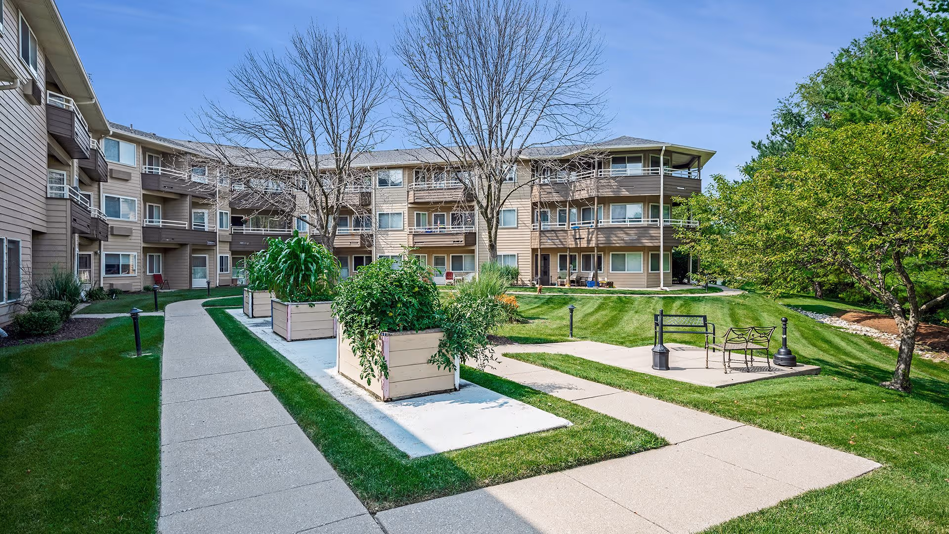 Outdoor courtyard area of a senior living facility with a paved walkway, raised garden beds with plants, benches, and a three-story building in the background under a clear blue sky.
