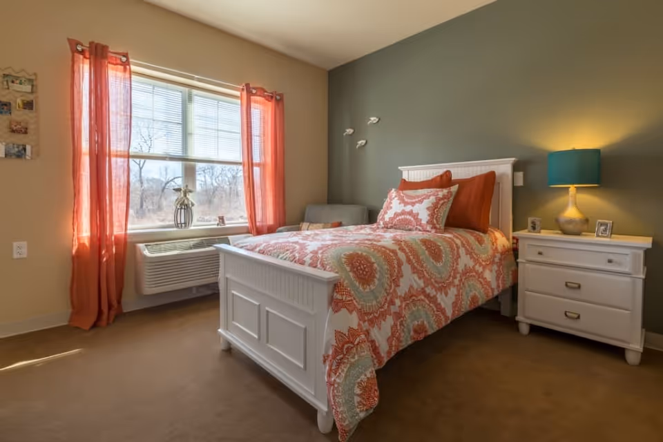 A cozy bedroom with a white wooden bed frame and a colorful patterned bedspread in shades of orange, green, and white. There are two pillows on the bed, one matching the bedspread and the other solid orange. Next to the bed is a white nightstand with two drawers, a teal lamp, and two small framed photos. The walls are painted in two tones, beige and green. A window with orange curtains lets in natural light, and below the window is an air conditioning unit. A small gray chair is positioned in the corner near the bed.