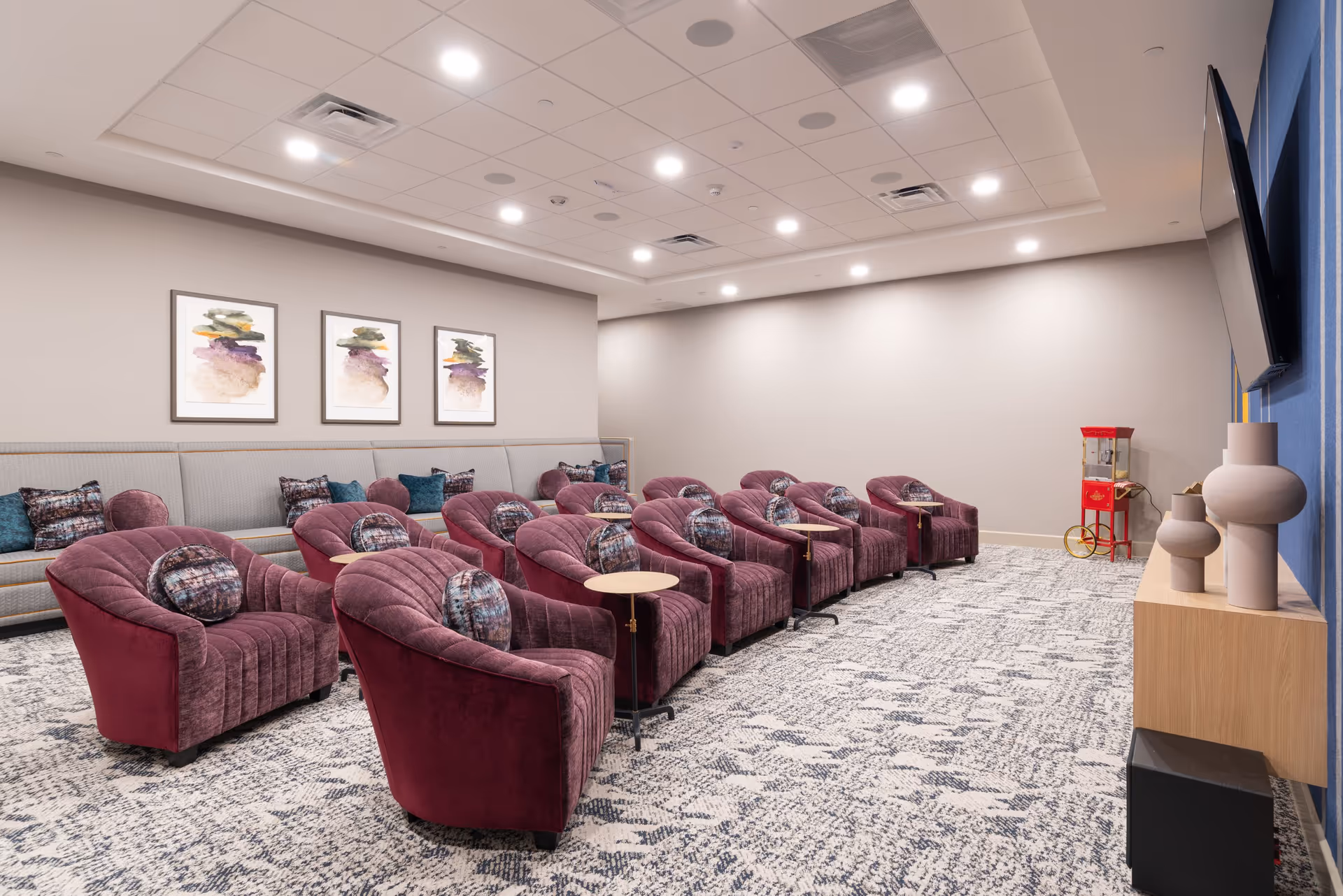 A modern lounge or media room with two rows of plush purple armchairs, each with a small attached table and decorative pillows. Behind the chairs is a long gray bench with additional pillows. The walls are light gray with three abstract paintings hanging above the bench. On the right side, a large flat-screen TV is mounted on a blue accent wall above a wooden shelf holding decorative vases. A red popcorn machine is visible in the back corner of the room. The floor is covered with a patterned carpet.