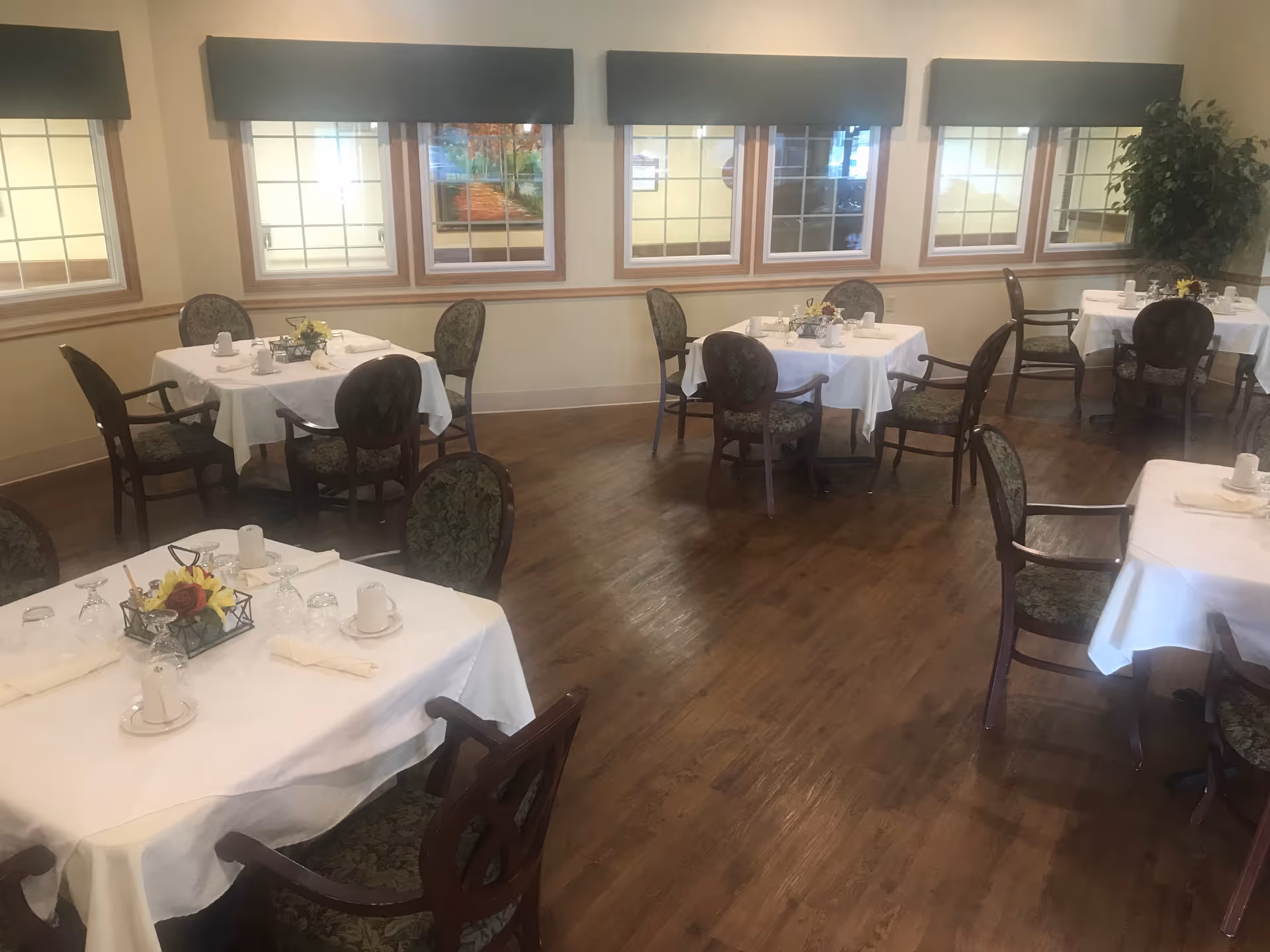 A dining room with several tables covered in white tablecloths, each set with cups, glasses, napkins, and small floral centerpieces. The room has wooden floors, multiple windows with valances, and upholstered chairs around the tables.