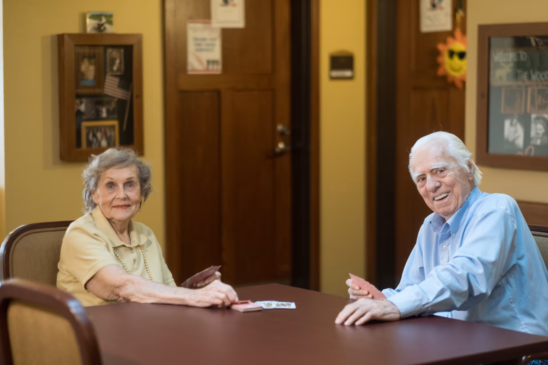 Two elderly individuals sitting at a table playing cards in a warmly lit room with wooden doors and framed pictures on the walls.