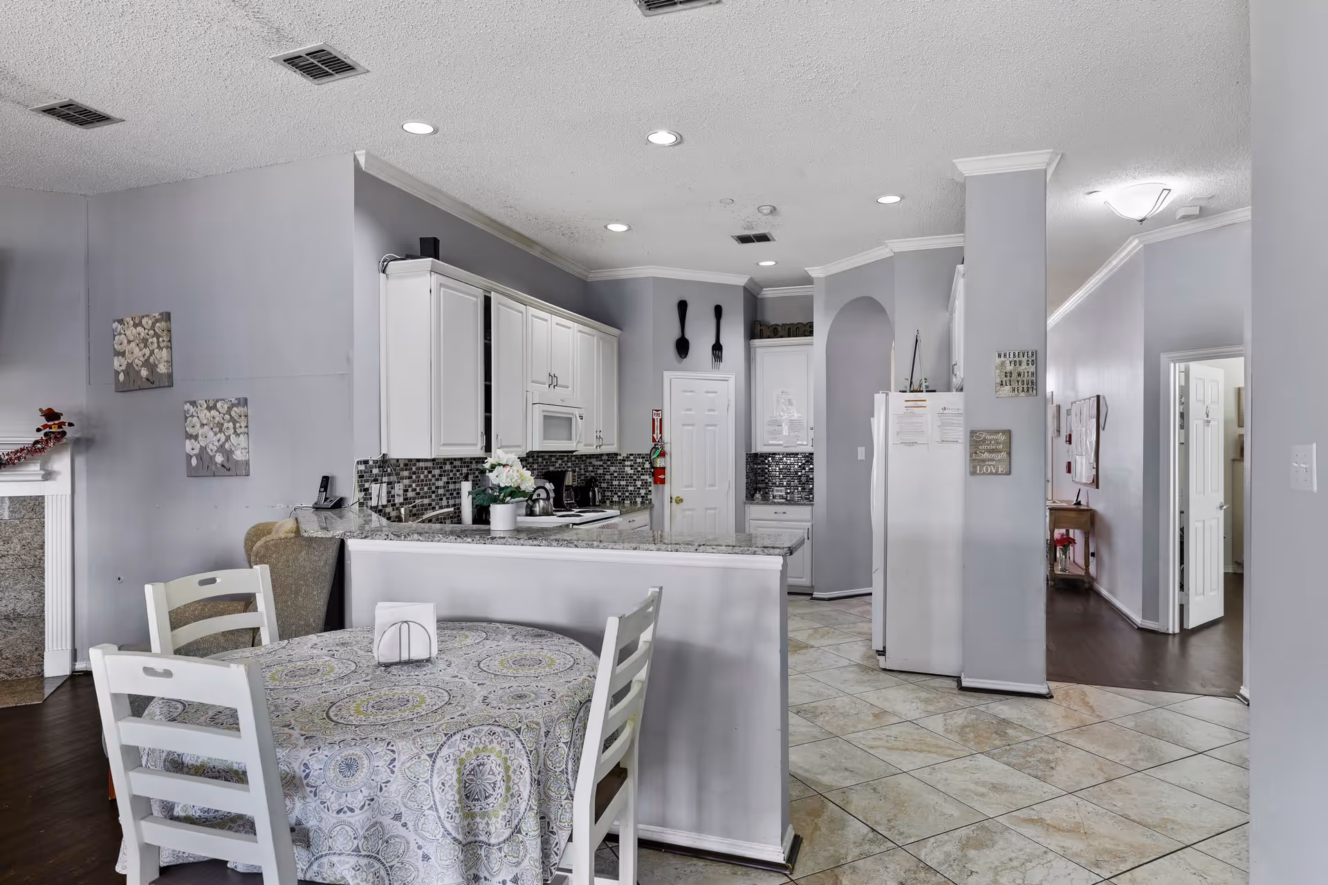 Interior view of a senior living facility kitchen and dining area. The kitchen features white cabinets, a tiled backsplash, and granite countertops. A round dining table with a patterned tablecloth and four white chairs is in the foreground. The floor is tiled, and the walls are painted light gray. Decorative wall art and kitchen utensils are visible, along with a refrigerator and a hallway leading to other rooms.