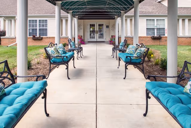 Covered outdoor walkway with black metal benches featuring teal cushions and decorative pillows on either side, leading to a building entrance with double glass doors and windows.