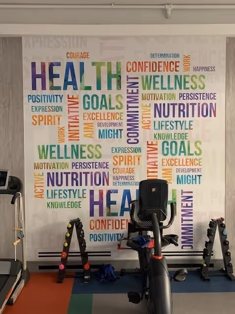 Indoor fitness area with exercise equipment including a stationary bike and dumbbell racks. The wall behind features a colorful word collage with terms related to health and wellness such as HEALTH, WELLNESS, NUTRITION, GOALS, COMMITMENT, and CONFIDENCE.