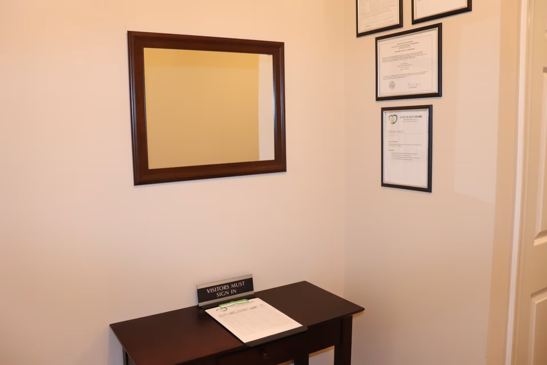 A small wooden table with a sign that reads 'VISITORS MUST SIGN IN' and a clipboard with a sign-in sheet. Above the table is a rectangular mirror with a dark wooden frame. On the adjacent wall, there are three framed certificates or documents, one of which has the logo and name 'Kind Hearts Home Assisted Living, LLC'.