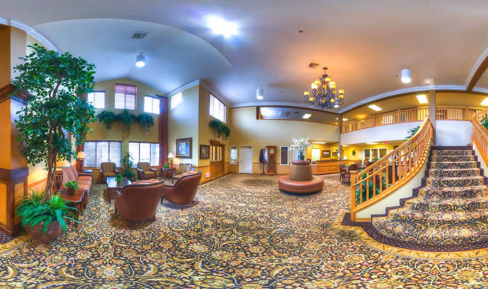 Spacious senior living facility common area with patterned carpet, comfortable seating including armchairs and sofas, large potted plants, and a staircase with wooden railings leading to an upper floor. The room is well-lit with ceiling lights and a chandelier, and has large windows with curtains allowing natural light.