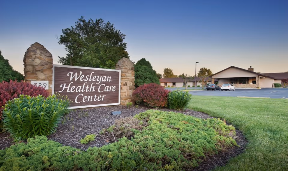 A landscaped entrance sign reading 'Wesleyan Health Care Center' with the facility building and parking lot visible behind it.