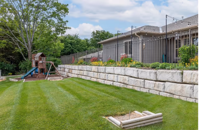 Outdoor area with a well-maintained green lawn, a wooden playset with a slide and swings, a stone retaining wall with flowers and shrubs planted on top, and a building with a gray exterior and string lights above the patio area under a partly cloudy sky.