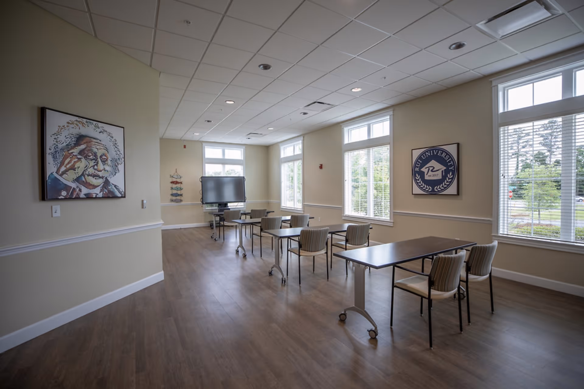 A bright, spacious room with several tables and chairs arranged in rows facing a large screen on a stand. The room has large windows with white blinds letting in natural light, light-colored walls, and wood flooring. On the walls are two framed pictures, one of Albert Einstein and another with a logo that reads 'RUI University est. 2017'.