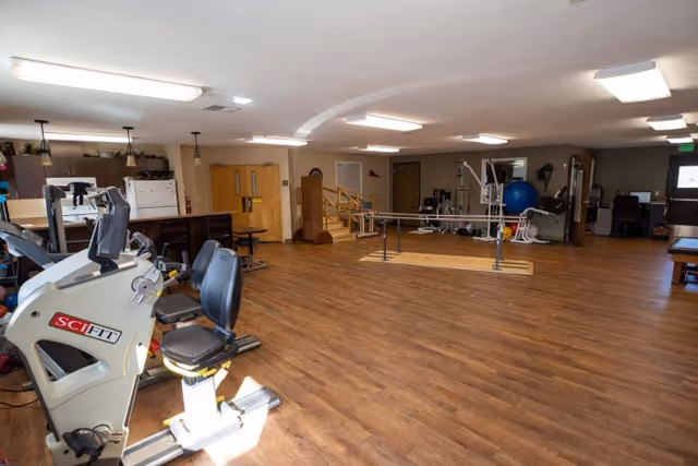 A spacious rehabilitation or physical therapy room with wooden flooring, exercise equipment including a recumbent exercise bike, parallel bars for walking practice, a large blue exercise ball, and various therapy tools. The room has bright overhead lighting and a small kitchenette area with a refrigerator and microwave in the background.