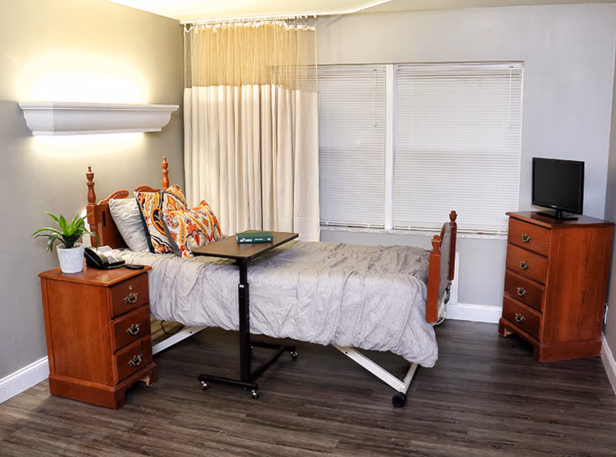 A bedroom in a senior living facility with a single bed featuring a gray comforter and colorful pillows. There is a wooden nightstand with a potted plant and a telephone on the left side of the bed. A small overbed table is positioned over the bed. On the right side of the room, there is a wooden dresser with a small flat-screen TV on top. The room has wood flooring, gray walls, a wall-mounted light fixture, and two windows with closed blinds partially covered by a beige curtain.