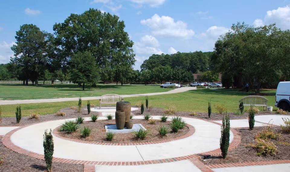 Outdoor garden area with a circular paved walkway surrounding a central water fountain feature made of three stacked stone pots. There are small plants and shrubs planted around the walkway, two wooden benches facing the fountain, a grassy field with large trees in the background, several parked cars, and a partly cloudy blue sky.