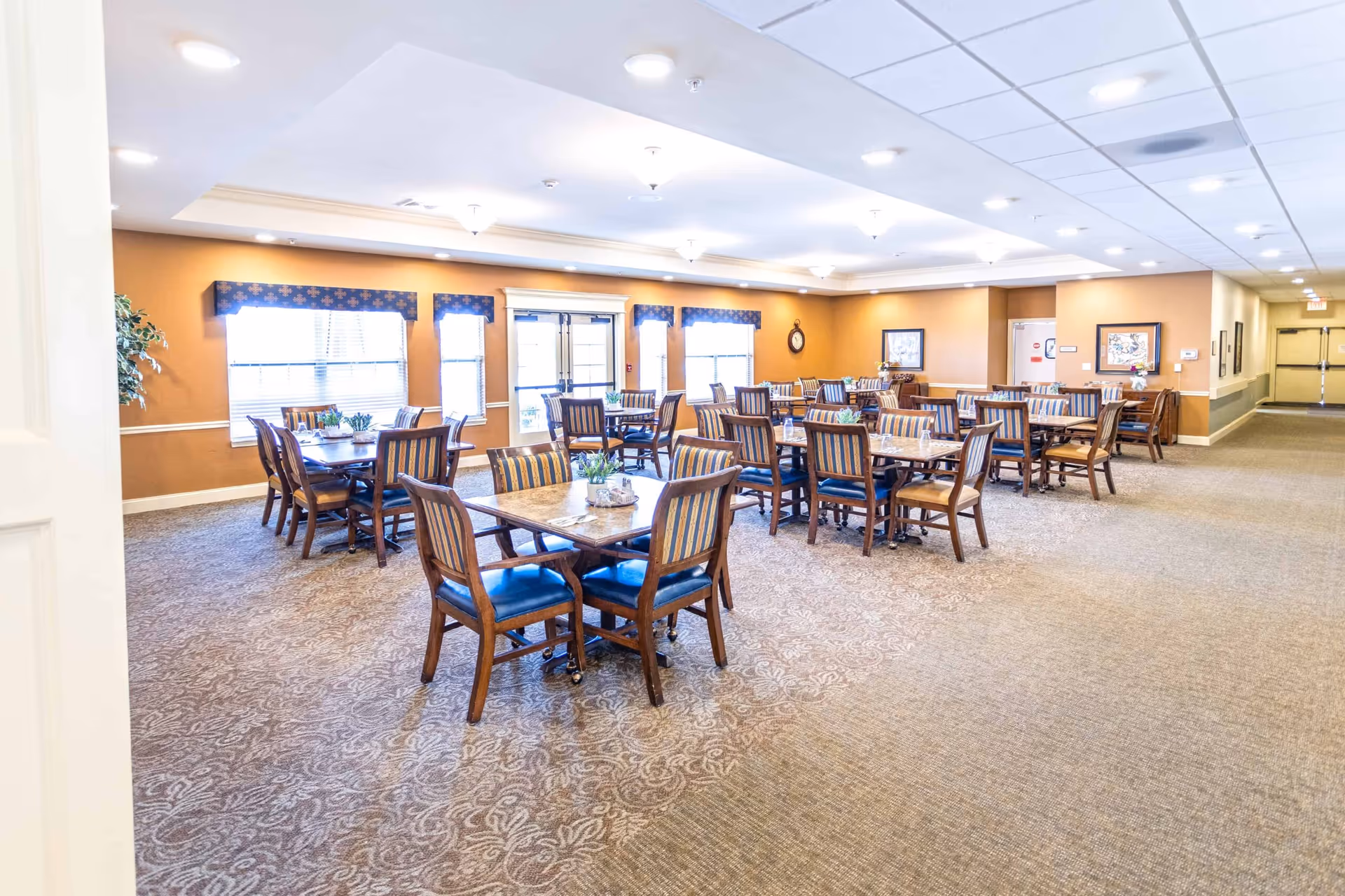 A spacious dining room with multiple wooden tables and chairs arranged neatly. The room has beige walls with white trim, patterned carpet flooring, and several windows with blue valances allowing natural light to enter. The ceiling features recessed lighting and decorative light fixtures. There are framed pictures and a clock on the walls, and a hallway is visible on the right side.