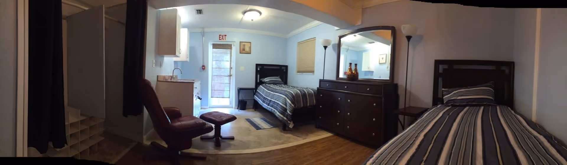 A cozy bedroom with two single beds covered in blue and white striped bedding. Between the beds is a dark wooden dresser with a large mirror and decorative vases on top. A floor lamp stands next to the dresser. On the left side of the room, there is a red leather recliner chair with a matching footrest, a small kitchenette with a sink, and a door with an exit sign above it. The walls are painted light blue and the floor is a combination of carpet and wood.