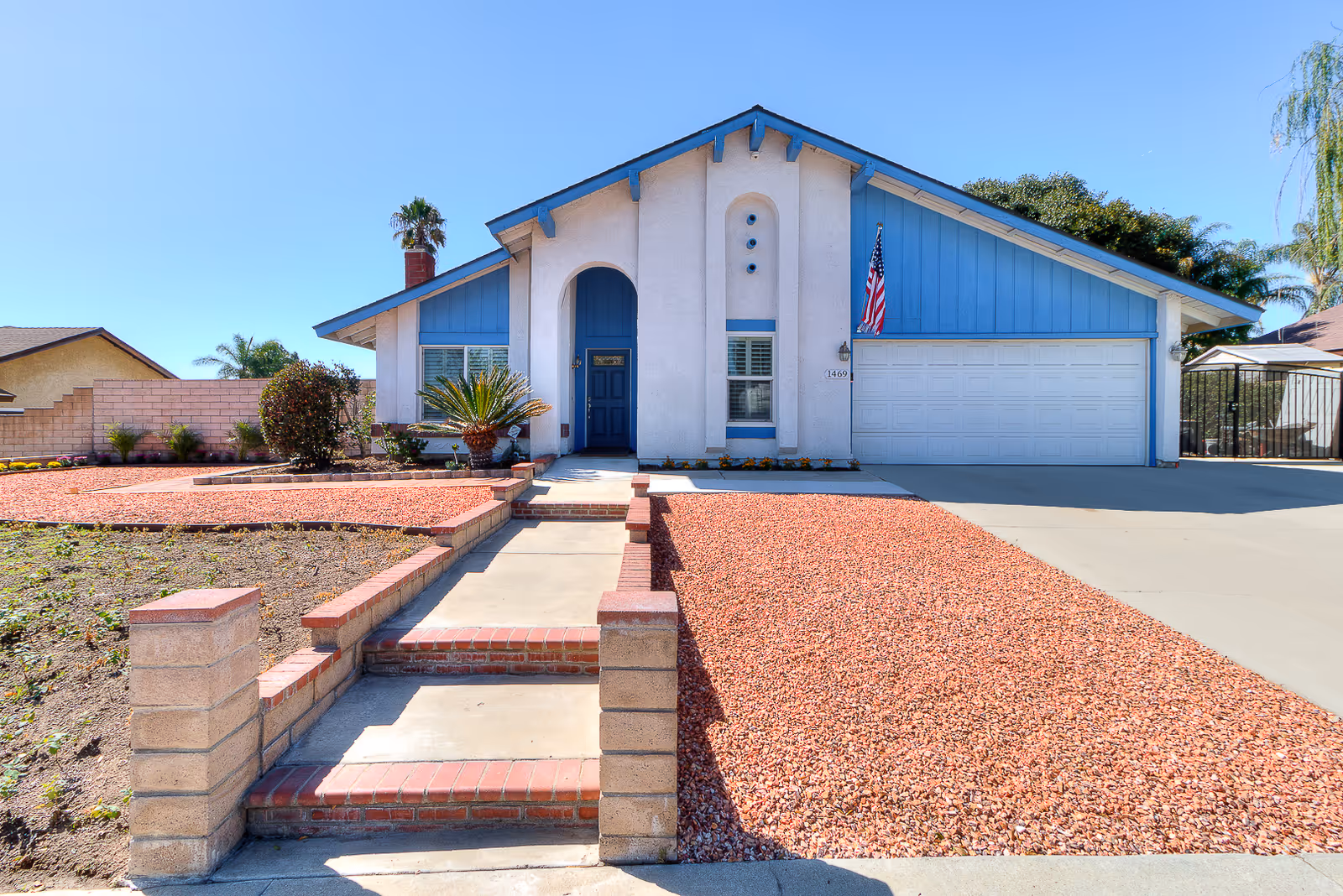 Front exterior view of a single-story house with a blue and white facade, a blue front door, a two-car garage, and a landscaped front yard with red gravel and a concrete walkway leading to the entrance.