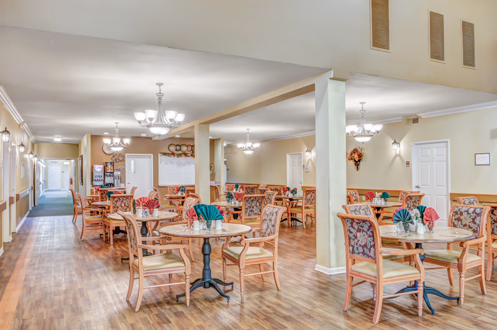 A spacious dining room in an assisted living facility with multiple round wooden tables and cushioned chairs featuring floral upholstery. Each table is set with folded red and green napkins, salt and pepper shakers, and small flower vases. The room has warm beige walls, wooden flooring, and several ceiling light fixtures providing bright illumination. A hallway extends from the dining area with doors along its length.