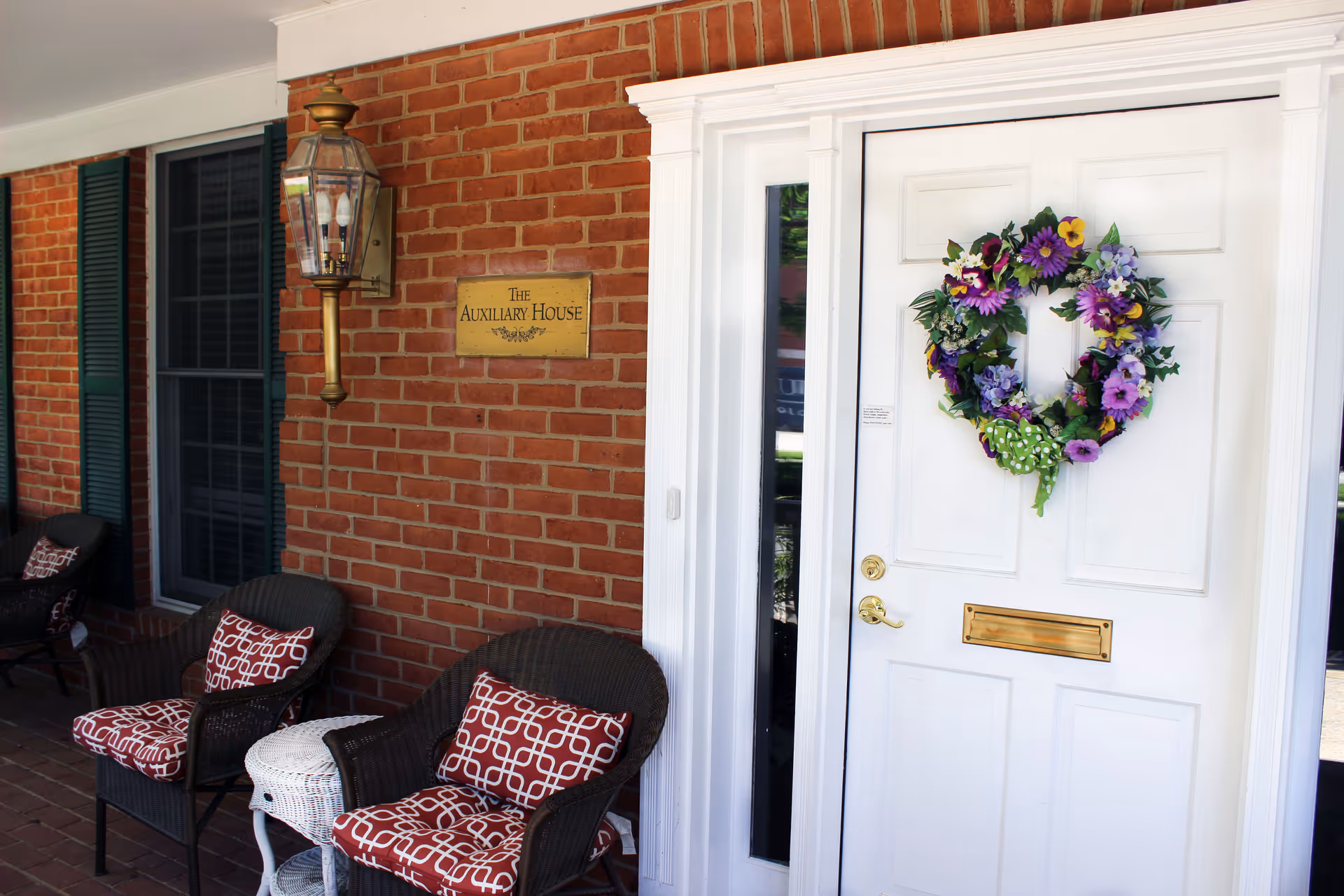 Front porch of a brick house showing a white door with a purple floral wreath, a brass plaque reading 'The Auxiliary House', and wicker chairs with red patterned cushions.