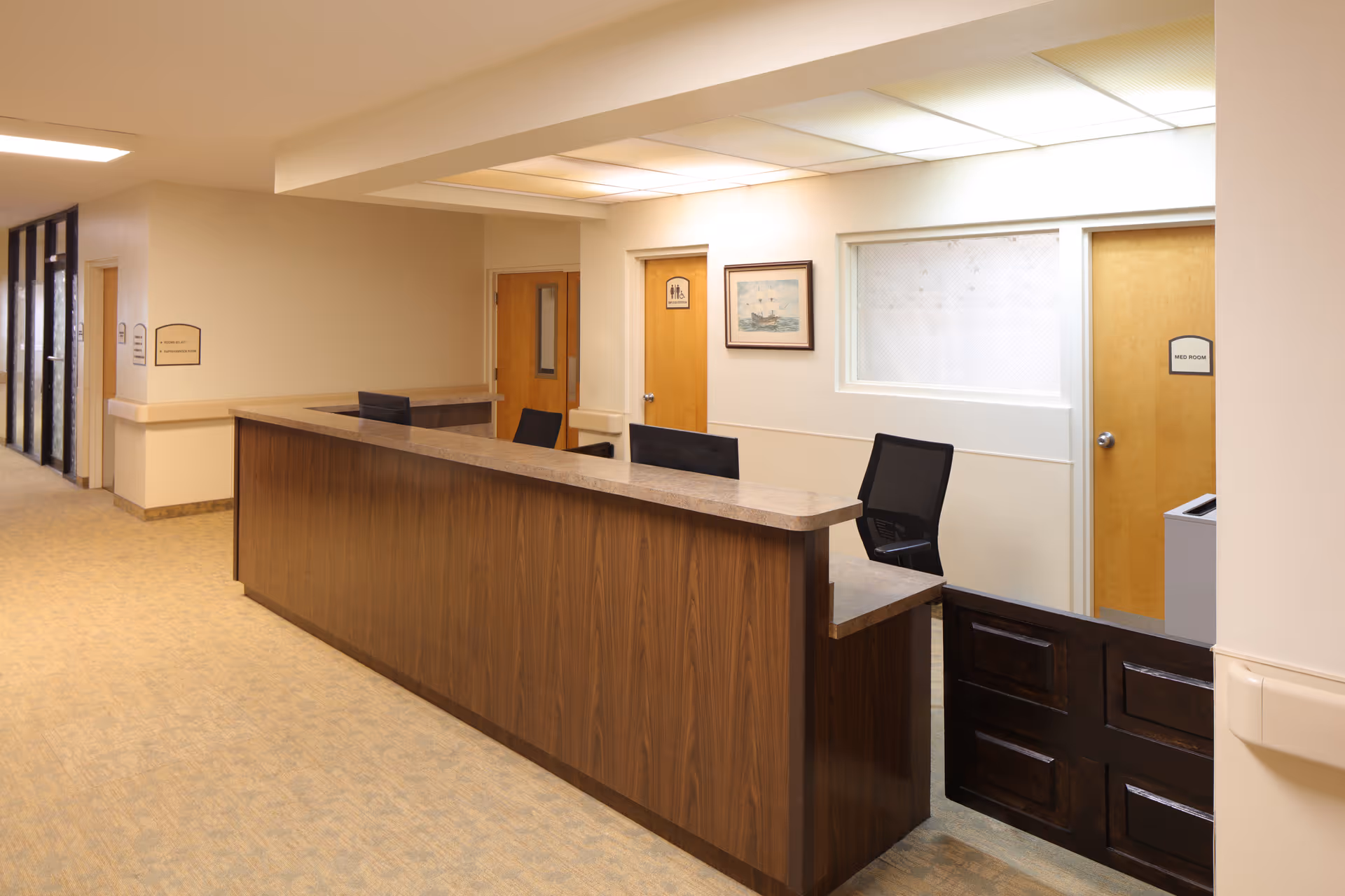 Reception desk in a facility hallway with chairs, doors, and framed artwork on the wall.
