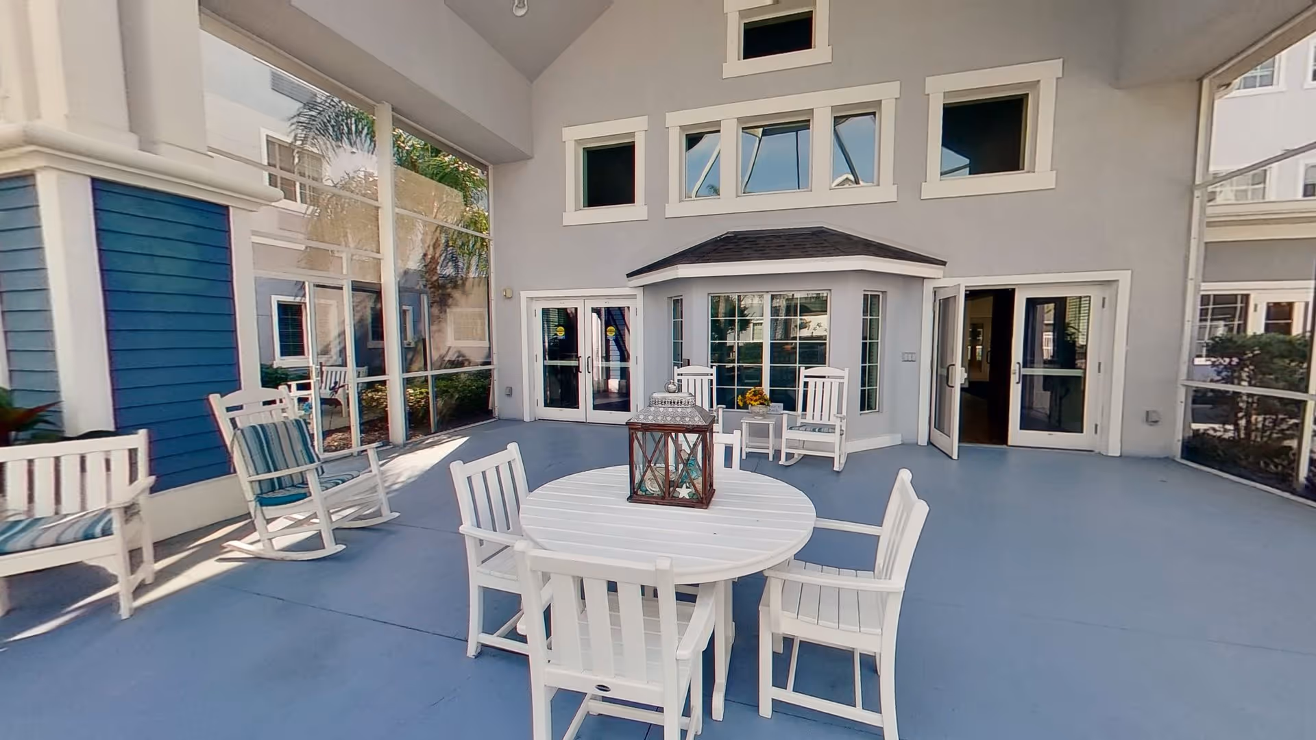 Covered outdoor patio area with white wooden chairs and a round table with a decorative lantern on top. The patio is adjacent to a building with large windows and glass doors, and there are rocking chairs with striped cushions along the side.