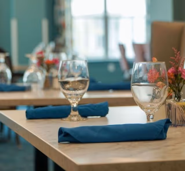 Close-up of a dining table set with wine glasses, blue cloth napkins, and small flower arrangements in a bright dining room.