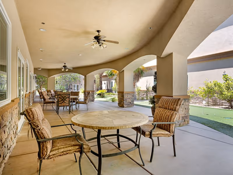 Covered outdoor patio area with multiple round tables and cushioned chairs. The patio has a beige ceiling with ceiling fans and is supported by stone and stucco columns. Beyond the patio, there is a landscaped garden area with green grass and shrubs.