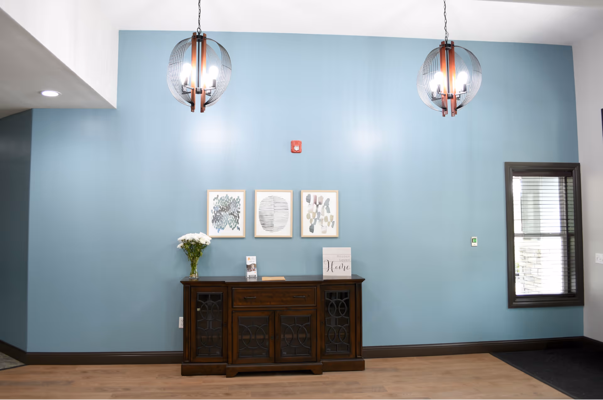 Interior view of a room with a light blue wall featuring three framed abstract art pieces. Below the artwork is a dark wooden cabinet with glass-paneled doors, decorated with a vase of white flowers and some small items. Two modern pendant lights hang from the ceiling, and there is a window with blinds on the right side.
