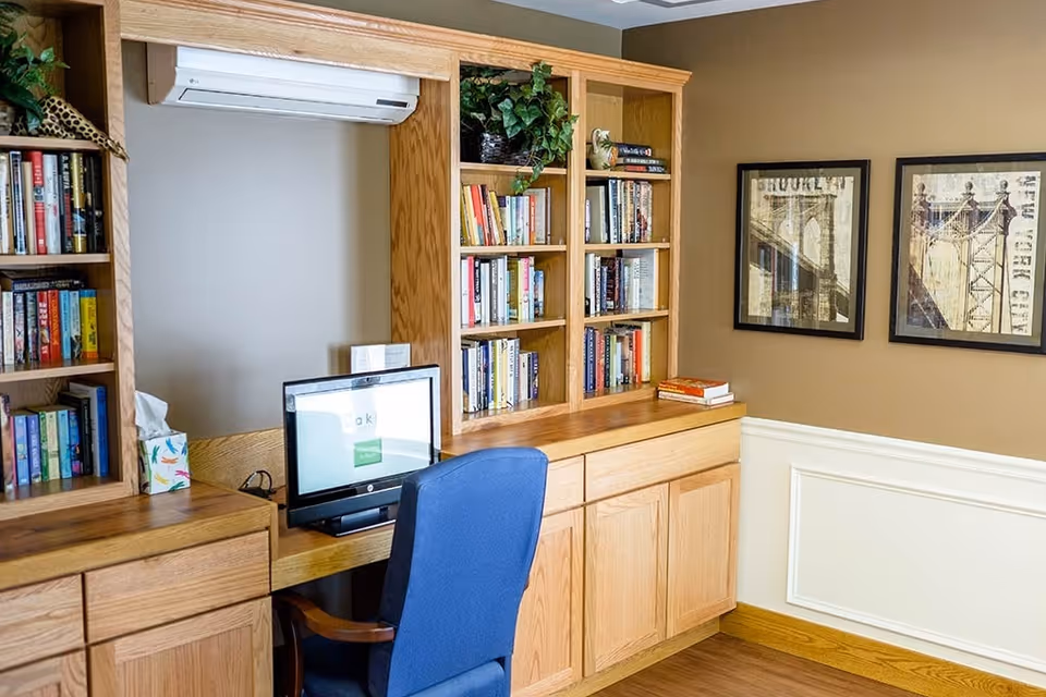A cozy office or study area with wooden built-in bookshelves filled with books and decorative plants. A computer monitor is on the desk with a blue office chair in front of it. The walls are painted beige with white wainscoting, and two framed pictures of bridges hang on the wall.