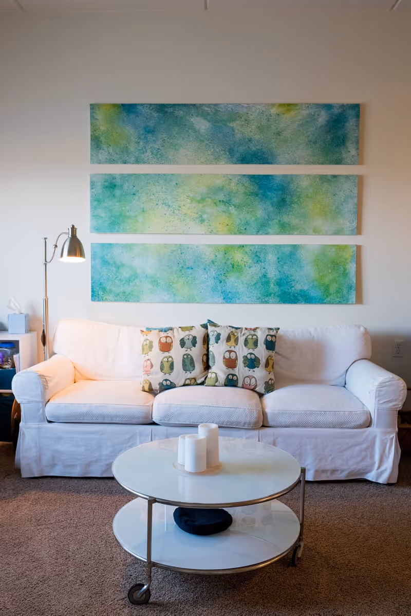 A cozy living room area with a white sofa adorned with three owl-patterned cushions. Above the sofa, there are three rectangular abstract paintings in shades of blue and green. A round white coffee table with two levels is placed in front of the sofa, holding three white candles on the top surface and a dark object on the lower shelf. A floor lamp with a metallic finish stands to the left of the sofa, next to a small side table with a tissue box and other items.