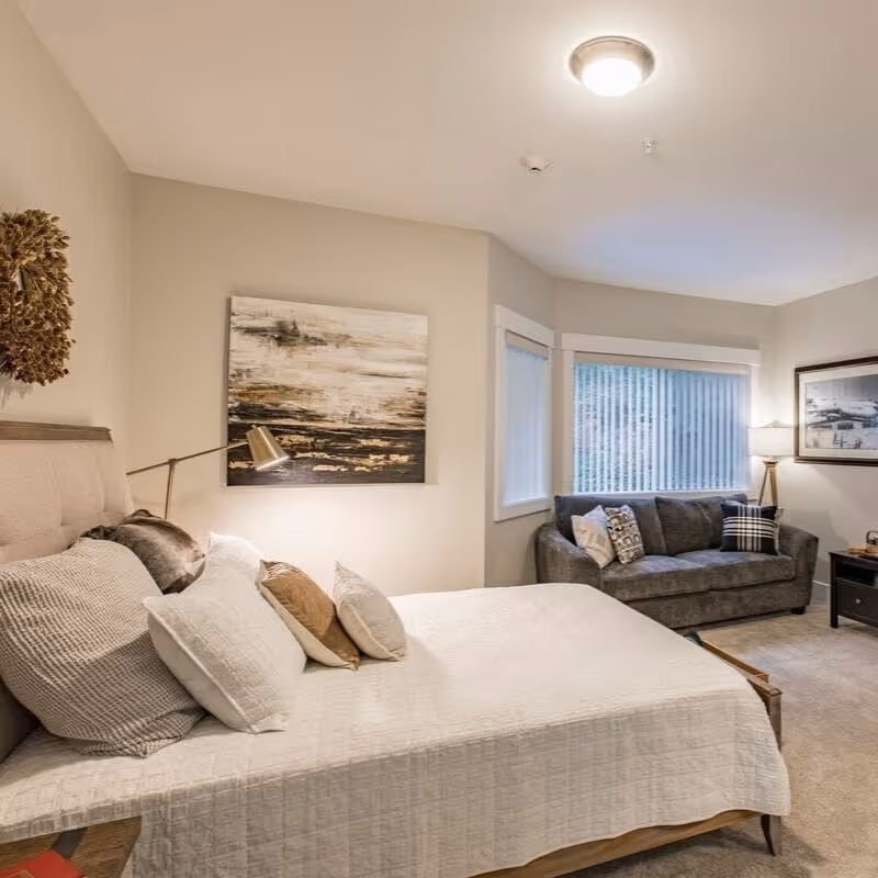 A cozy bedroom with a neatly made bed featuring multiple pillows and a white quilt. To the right, there is a gray sofa with decorative pillows positioned near two windows with vertical blinds. The room is softly lit by a ceiling light and a floor lamp next to the sofa. The walls are decorated with a large abstract painting above the bed and a framed picture near the sofa.