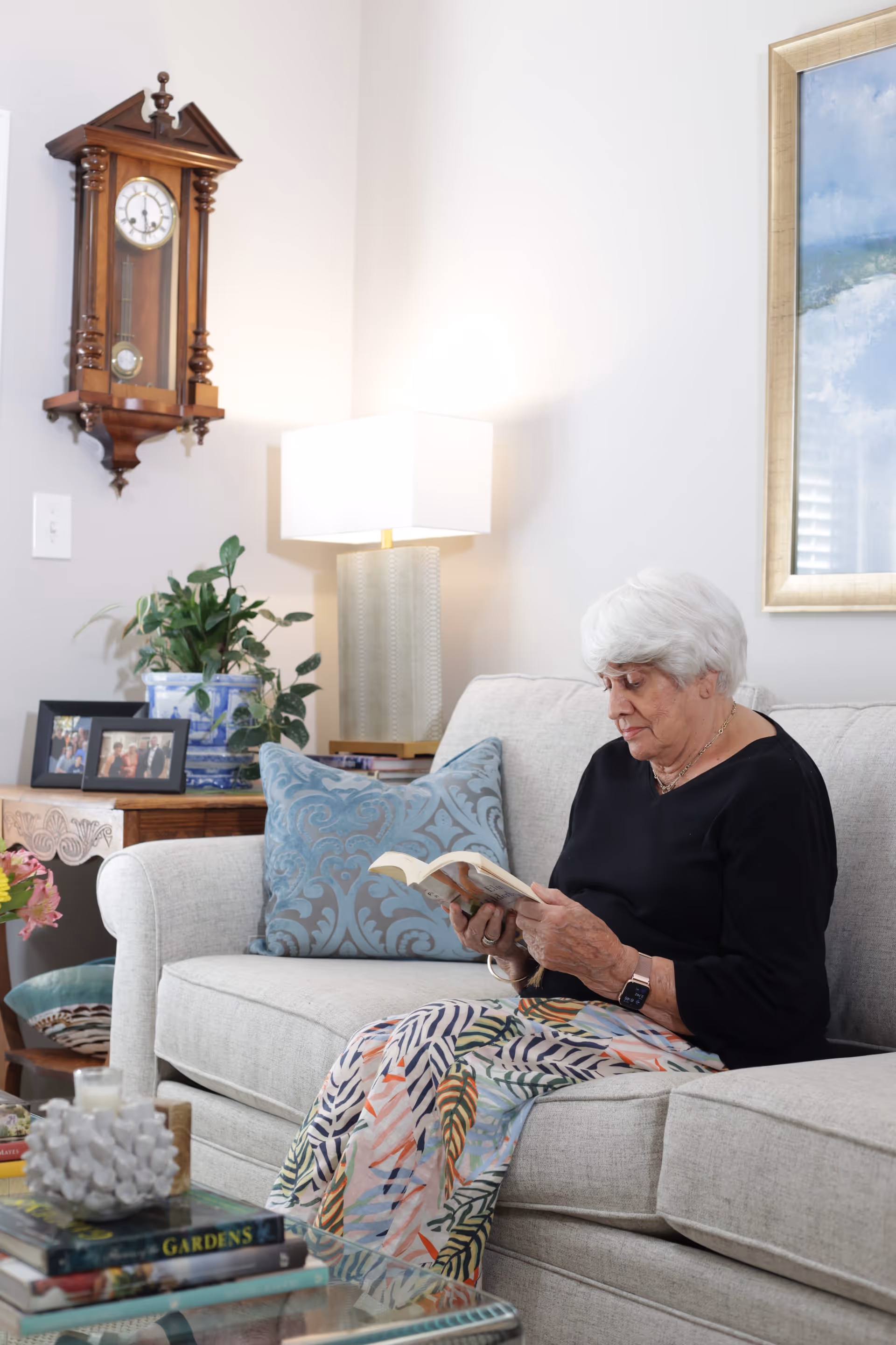 An elderly woman with white hair sits on a light gray sofa reading a book. She is wearing a black top and a colorful skirt with leaf patterns. Behind her is a wooden side table with framed photos, a potted plant, and a lamp with a white shade. A wooden wall clock hangs on the wall above the table, and a framed painting is partially visible on the right side of the image.