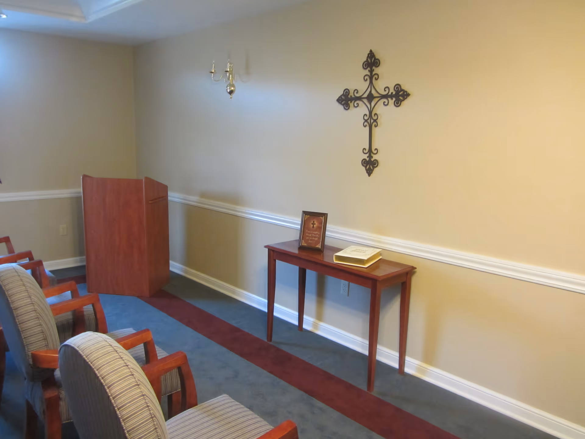A small room with several striped upholstered chairs arranged in rows facing a wooden podium. A narrow wooden table against the wall holds a framed picture and a book. A decorative metal cross hangs on the beige wall above the table. The room has a carpet with blue and maroon stripes and white chair rail molding on the walls.