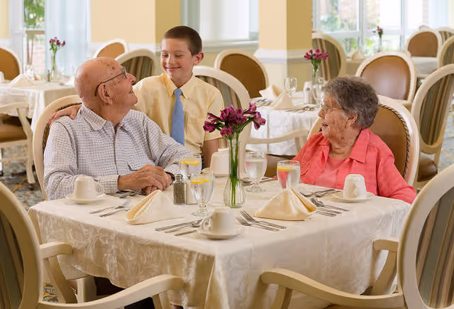 An elderly man and woman sit at a dining table in a well-lit dining room with white tablecloths and floral centerpieces. A young boy stands behind the man, smiling and placing his hand on the man's shoulder. The table is set with napkins, cups, glasses of water with lemon, and silverware.