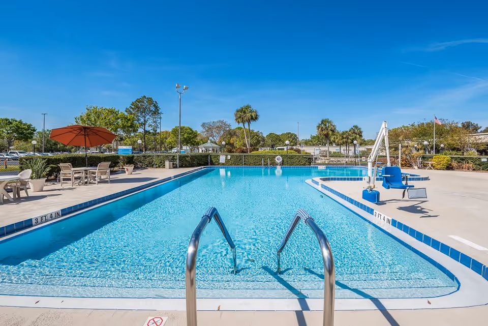 Outdoor swimming pool with lounge chairs, an umbrella, a pool lift, and palm trees under a clear blue sky.