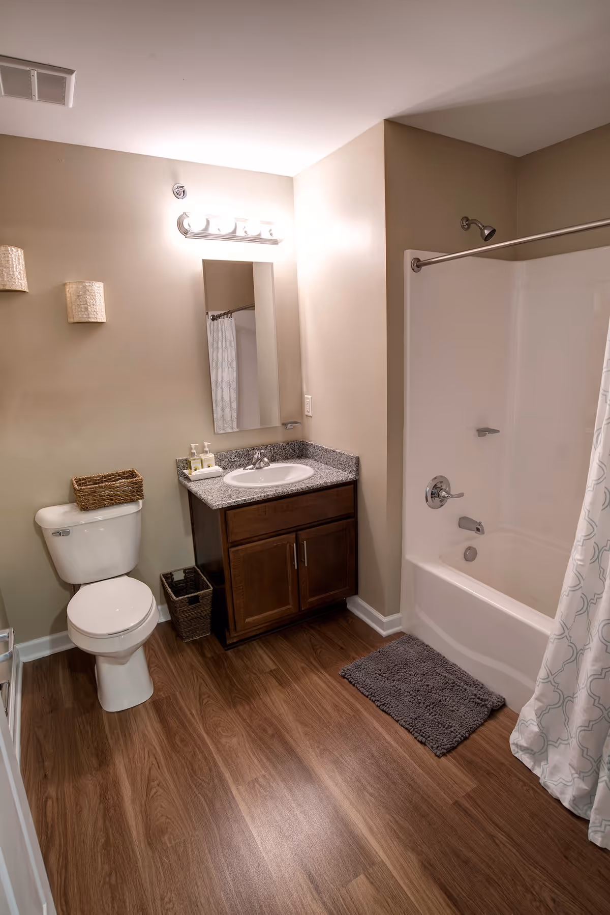 A clean bathroom with a white toilet, a wooden vanity with a granite countertop and a sink, a mirror above the sink, a bathtub with a showerhead and a white shower curtain with a subtle pattern, and a gray bath mat on the wooden floor.