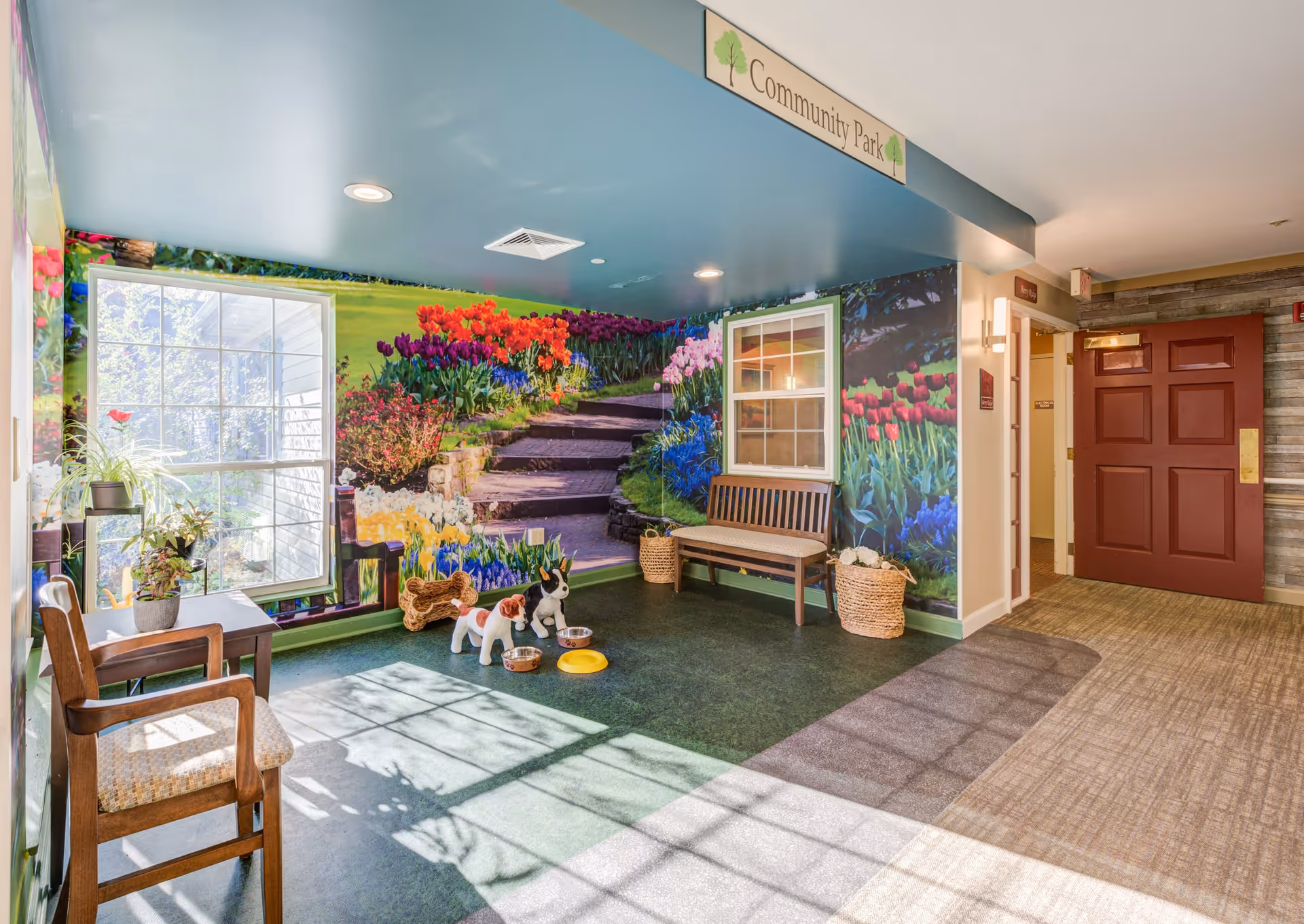 A bright indoor community space with a large window letting in natural light. The walls feature a colorful mural of a garden with flowers and stone steps. There are two wooden chairs, a bench, and some potted plants. On the floor, there are three dog statues with bowls. A sign above reads 'Community Park'.