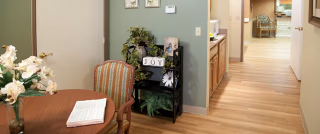 Interior view of a senior living facility hallway with light wood flooring and beige walls. In the foreground, there is a round wooden table with a striped upholstered chair and a vase of white flowers. A small black shelf with decorative items including a sign that says 'JOY' and some greenery is against a green accent wall. The hallway leads to another room with a chair and a desk visible in the background.