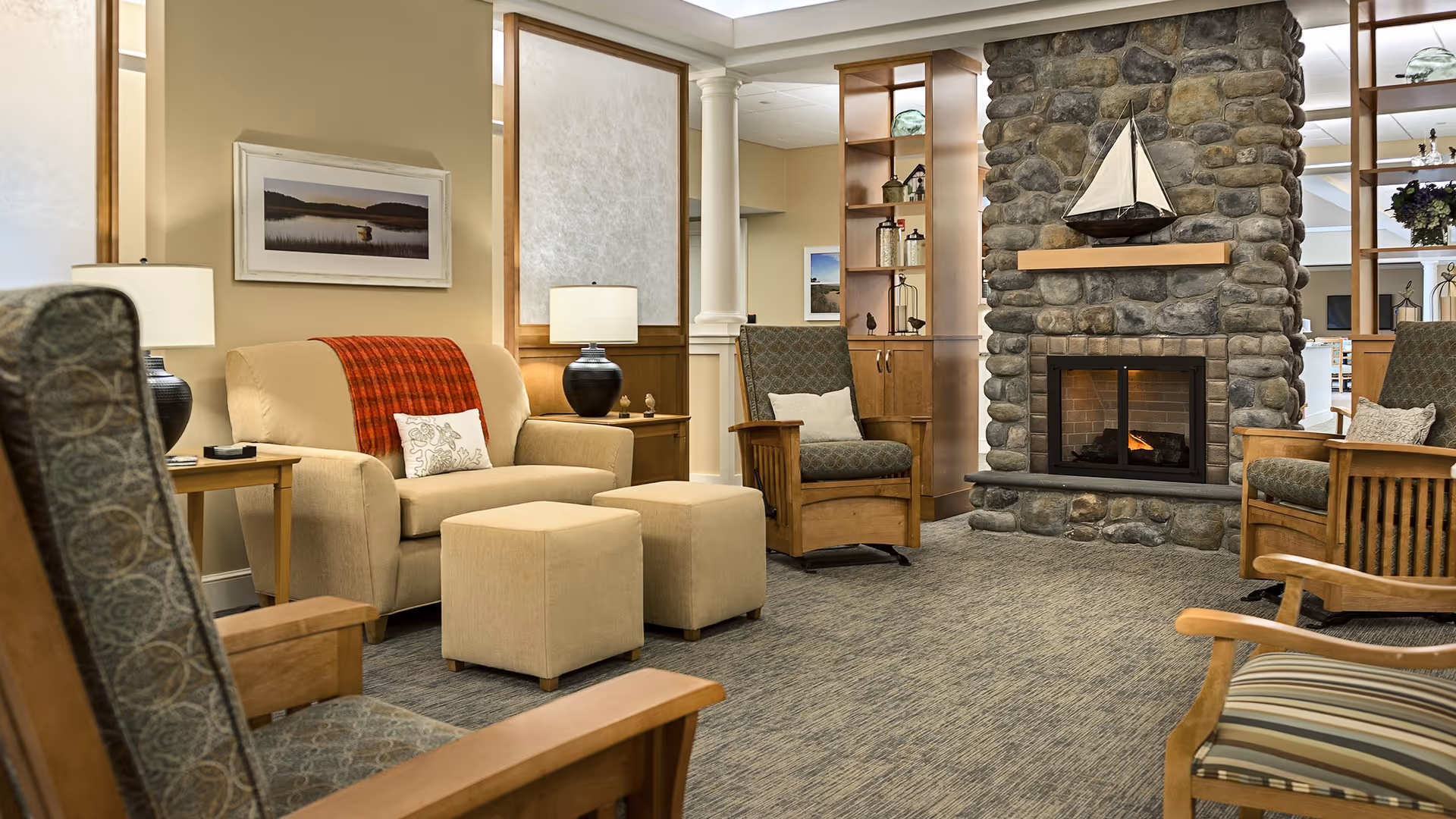 A cozy living room area in a senior living facility featuring a stone fireplace with a decorative sailboat on the mantel. The room has several wooden armchairs with patterned cushions, a beige loveseat with a red throw blanket and a decorative pillow, two beige ottomans, side tables with lamps, and built-in wooden shelving with decorative items. The walls are painted a warm beige color and there is framed artwork hanging on the walls.
