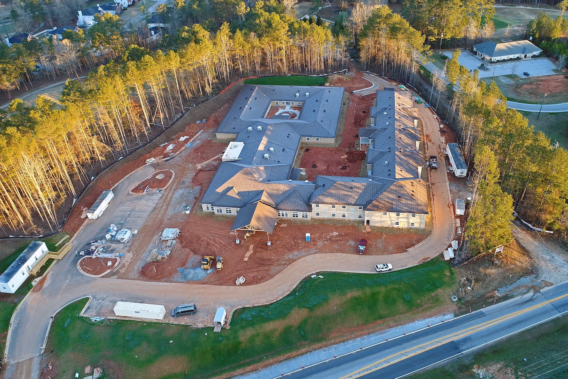 Aerial view of a large building under construction surrounded by trees and roads, with construction vehicles and materials visible around the site.