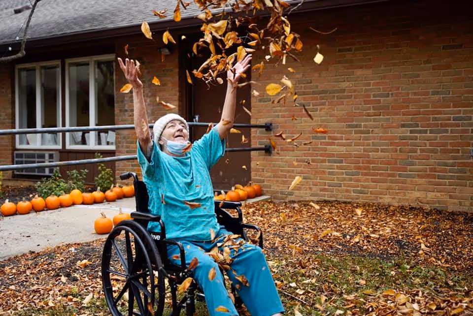 An elderly person in a wheelchair wearing a hospital gown and a knit hat joyfully throws autumn leaves into the air outside near a brick building with a row of pumpkins along the sidewalk.