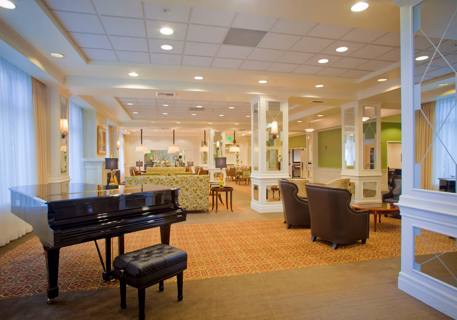 Spacious and well-lit common area in a senior living facility featuring a black grand piano with a matching padded bench in the foreground. The room has patterned carpet, multiple seating arrangements including floral sofas and leather armchairs, white columns with mirrors, and large windows with sheer curtains allowing natural light to fill the space.