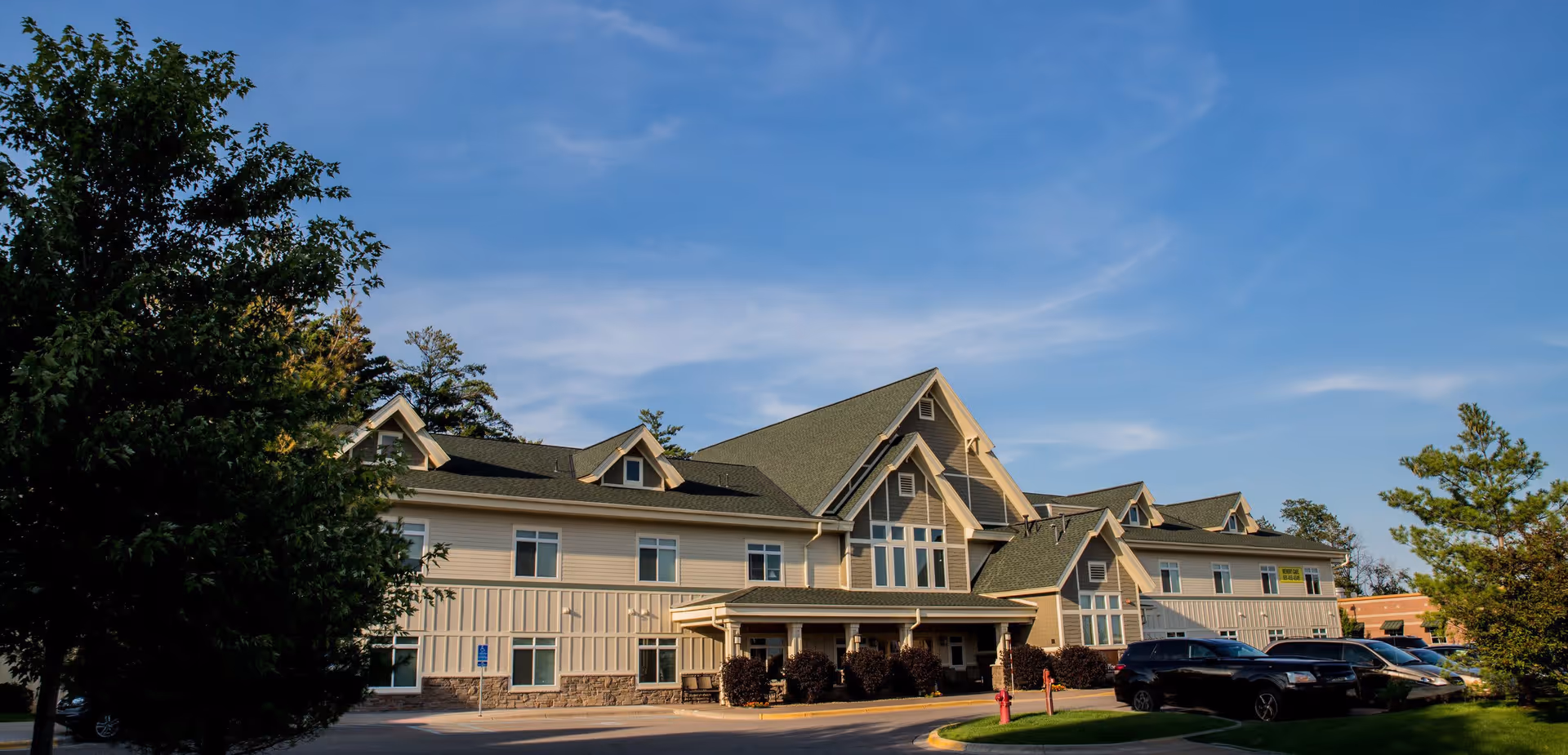 Exterior view of a two-story senior living facility building with beige siding, green roof, and multiple windows under a clear blue sky. There are trees and parked cars in front of the building.