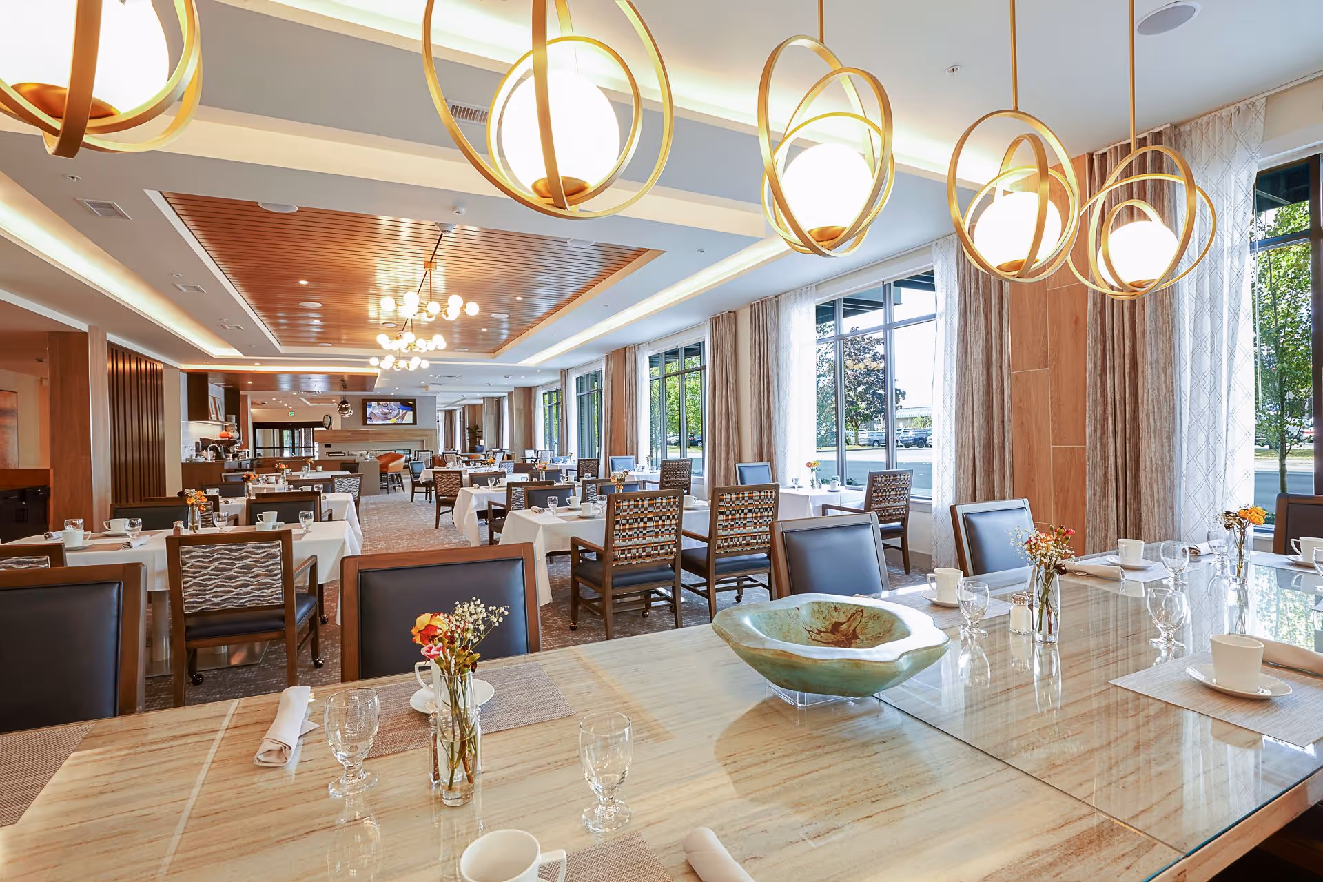 A bright and spacious dining room with multiple tables covered in white tablecloths, set with cups, glasses, napkins, and small flower arrangements. Large windows with curtains allow natural light to fill the room. Modern pendant lights hang from the ceiling, and a decorative bowl is placed on the foreground table.