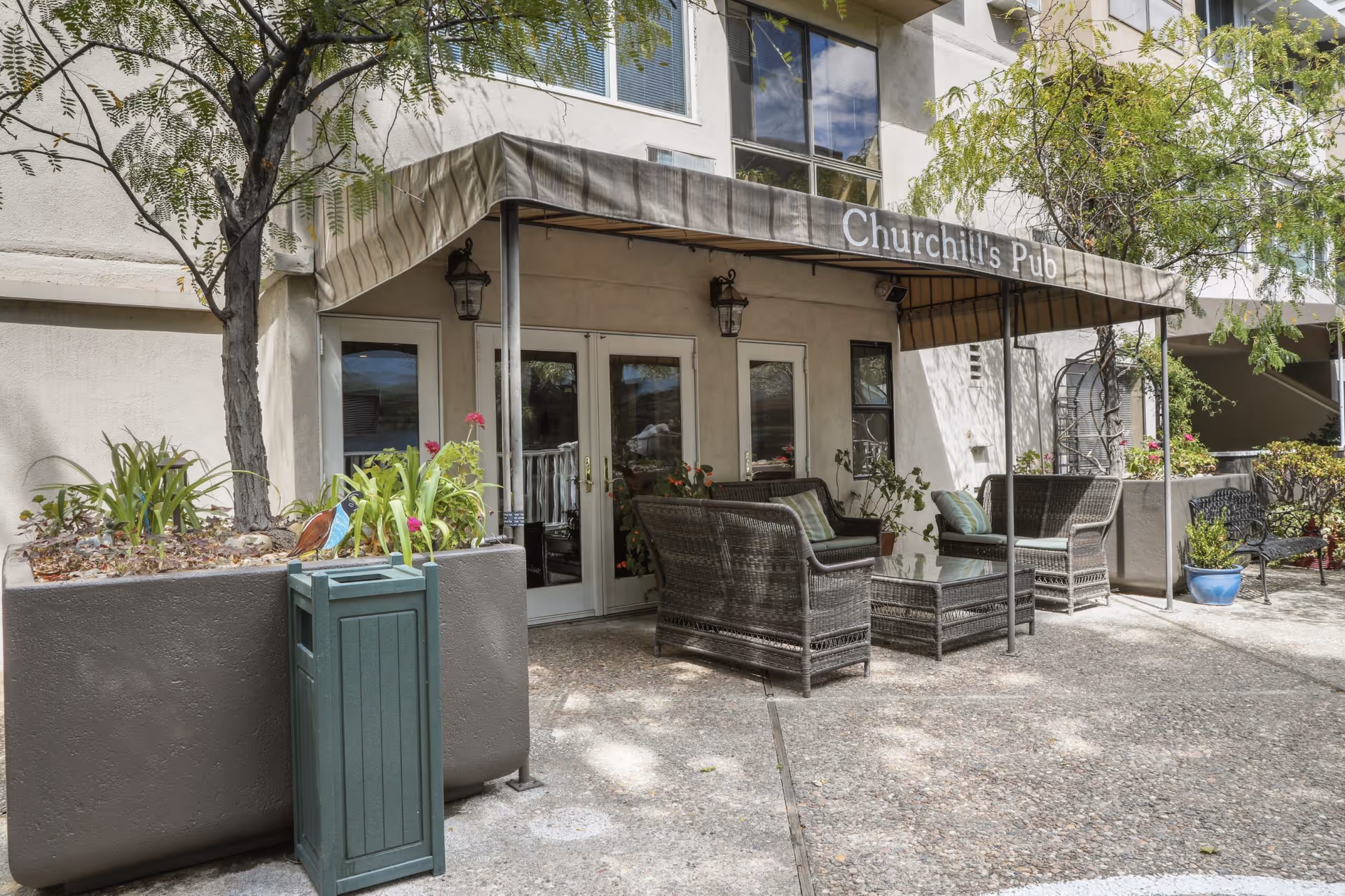 Outdoor patio area with wicker seating including chairs and a glass-top table under a canopy labeled 'Churchill's Pub'. There are potted plants and trees around the seating area, and the building exterior is visible in the background.