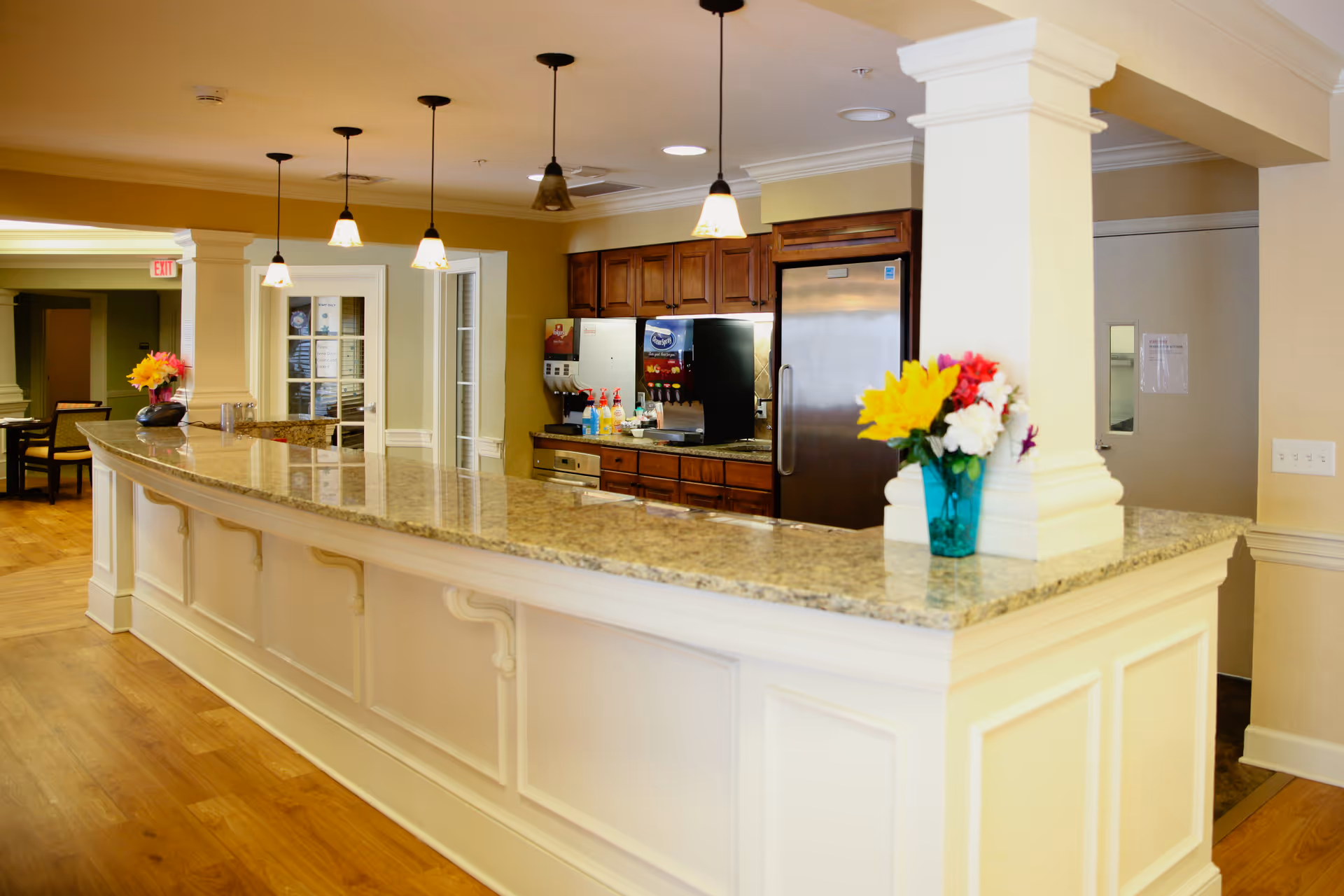 Interior view of a senior living facility's kitchen area with a long granite countertop bar, pendant lights hanging from the ceiling, wooden cabinets, a stainless steel refrigerator, and a beverage dispenser. There are colorful flower arrangements in vases on the countertop.