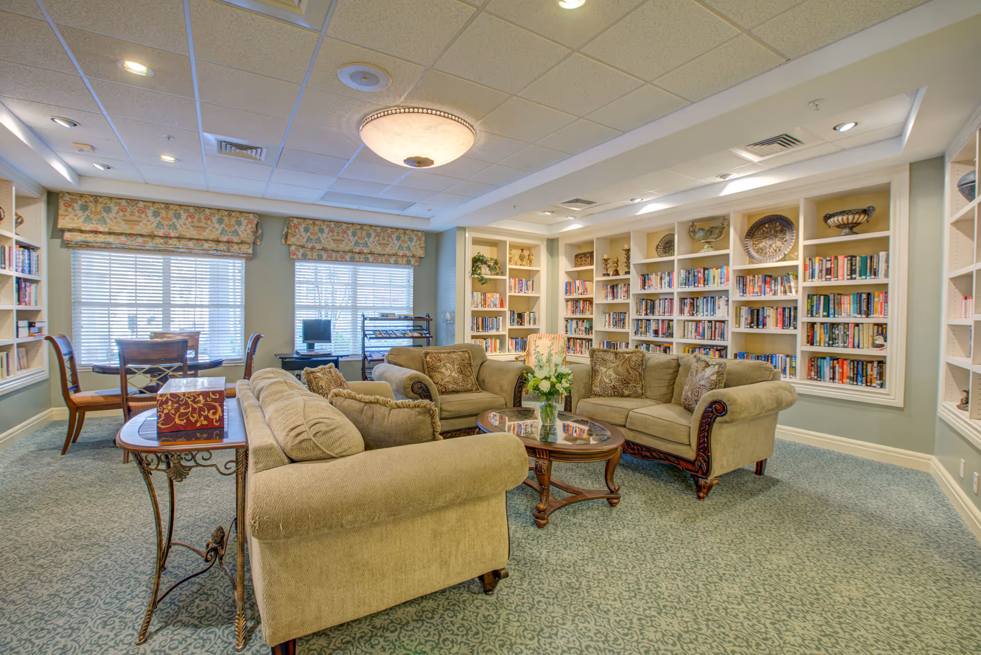 A cozy living room area with three beige upholstered sofas arranged around a wooden coffee table with a glass top and a vase of flowers. The room features built-in bookshelves filled with books and decorative items along the walls. There are two large windows with floral valances letting in natural light, a small table with chairs near the windows, and a computer workstation in the corner. The ceiling has recessed lighting and a central decorative light fixture, and the floor is carpeted with a patterned design.