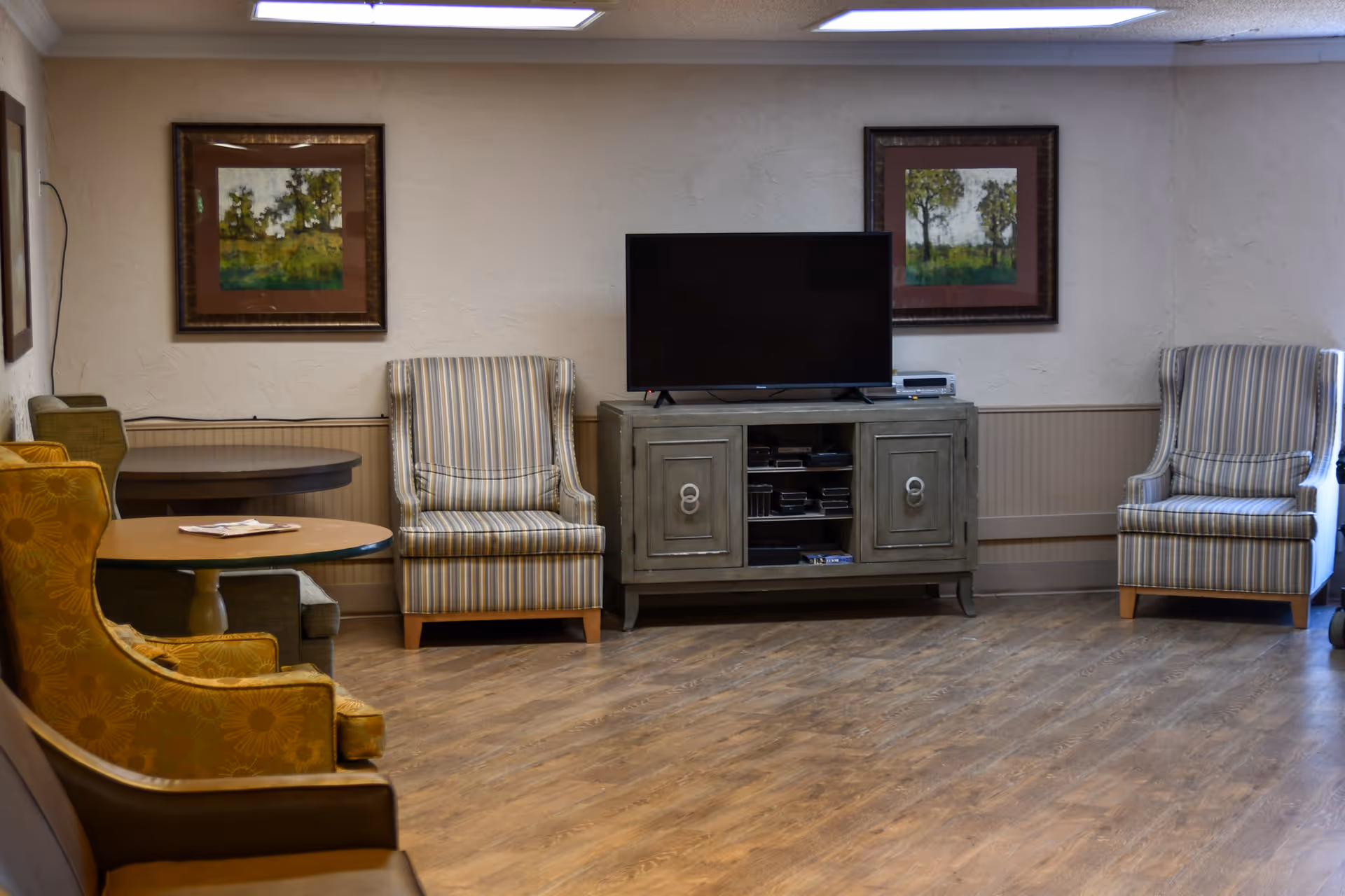 A cozy living room area with wooden flooring, featuring a TV on a gray cabinet, two striped armchairs on either side of the TV, a round wooden table with a yellow patterned armchair, and framed landscape paintings on the wall.