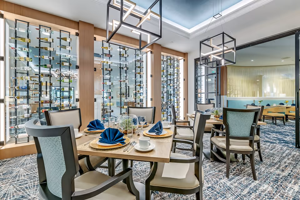 A modern dining room with a wooden table set for six people, featuring blue folded napkins, plates, cups, and glassware. The room has a patterned carpet and is illuminated by two rectangular pendant lights. Behind the table is a glass wall with multiple wine bottles displayed vertically on racks. In the background, there is another seating area with chairs and tables.