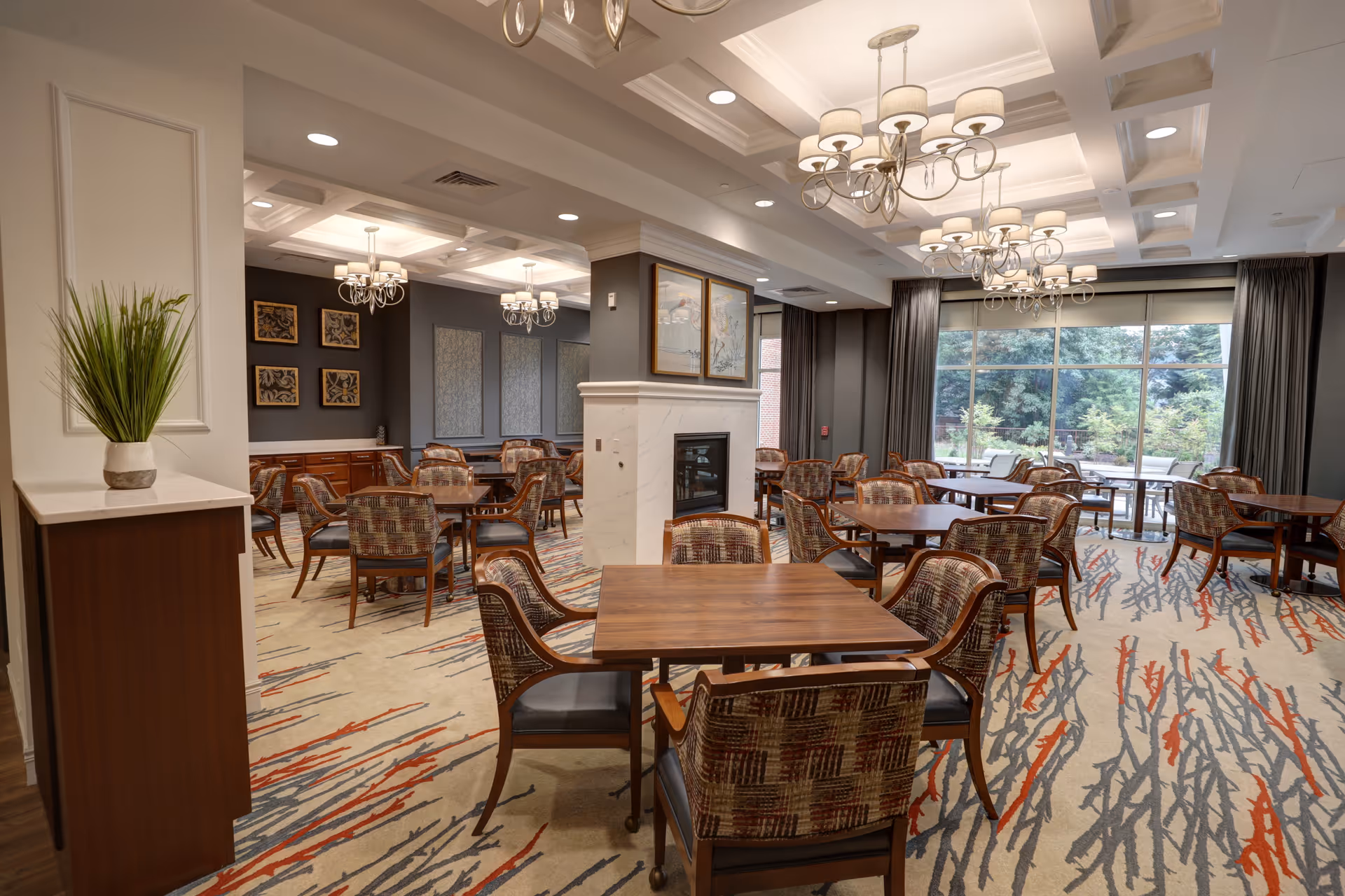 Bright communal dining room with multiple wooden tables and upholstered chairs, chandeliers, a central fireplace, and large windows overlooking greenery.